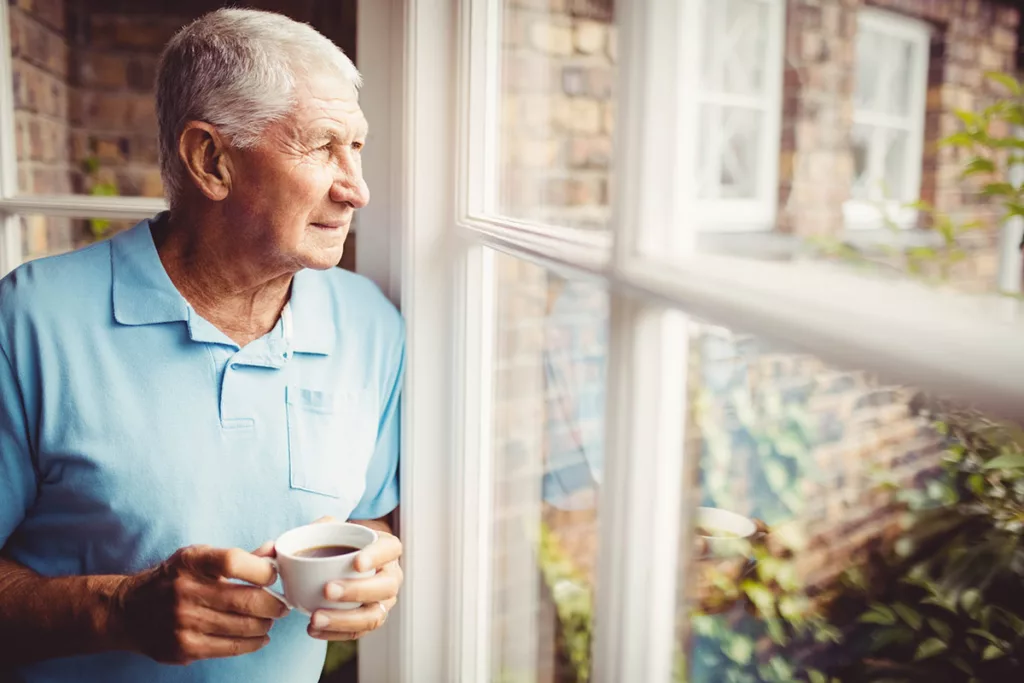 Man at home looking out window holding coffee cup.