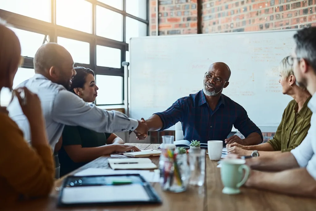 A. group of business colleagues, sitting in a conference room having a meeting, older business man shaking hands with co-worker, symbolizing how ai hearing aids help with speech clarity even in challenging listening environments.