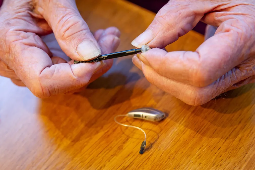 A man sitting at a kitten table holding a hearing aid wax pixk, demonstrating the concept of how to properly clean your hearing aids at home