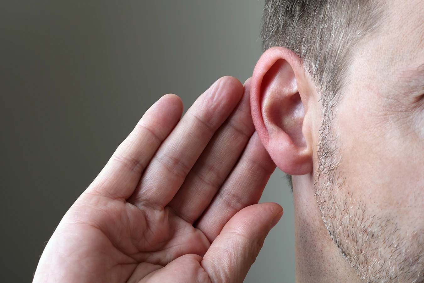 A mature man with grey and white, salt and pepper hair, holding his hand up to his ear in an effort to hear more clearly, symbolizing that better hearing often requires more than just turning up the volume.