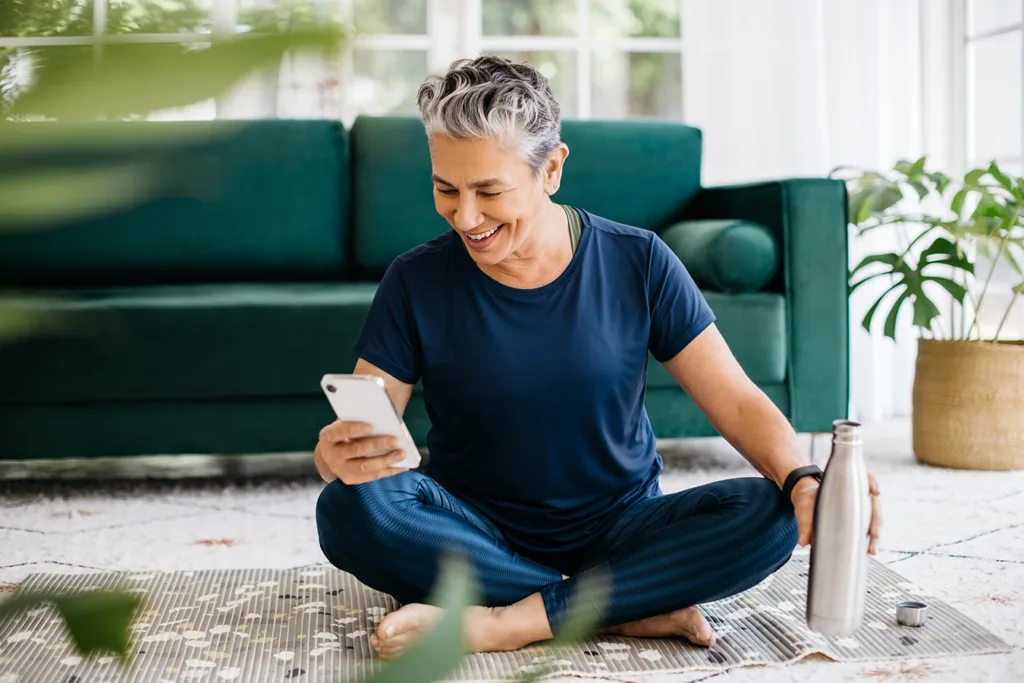 Happy, healthy, active senior woman using a smartphone after doing yoga in her living room, tracking her fitness progress on a smartphone app connected to her health tracking hearing aids. 